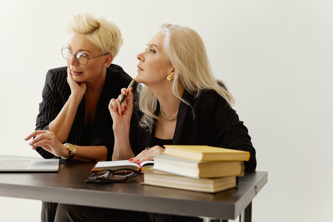 Two businesswomen in discussion, with books and a laptop on their desk, against a white background.