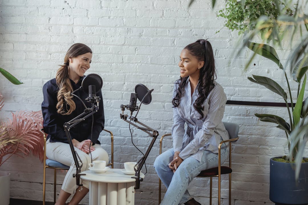 Two smiling women recording a podcast together in a room with white brick walls and plants.