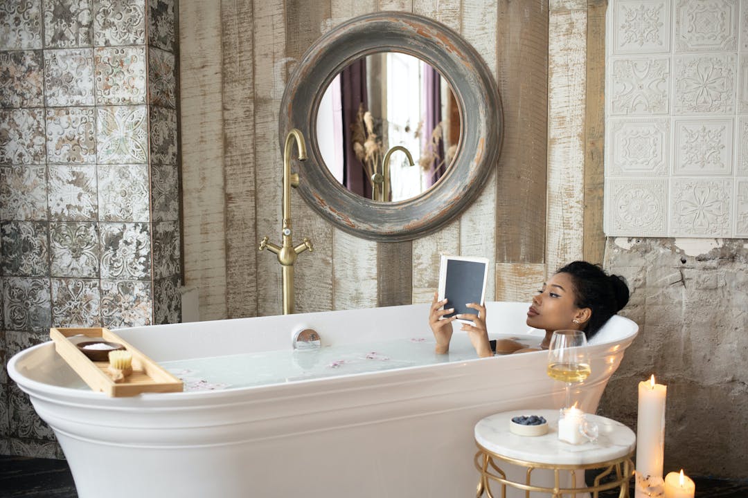 Woman relaxing in a bathtub, reading a tablet beside candles and a glass of wine